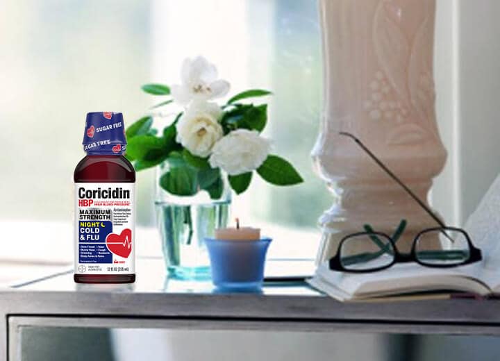 Bedroom nightstand with a bottle of Coricidin HBP Nighttime Multi-Symptom Cold Syrup, a book and glasses